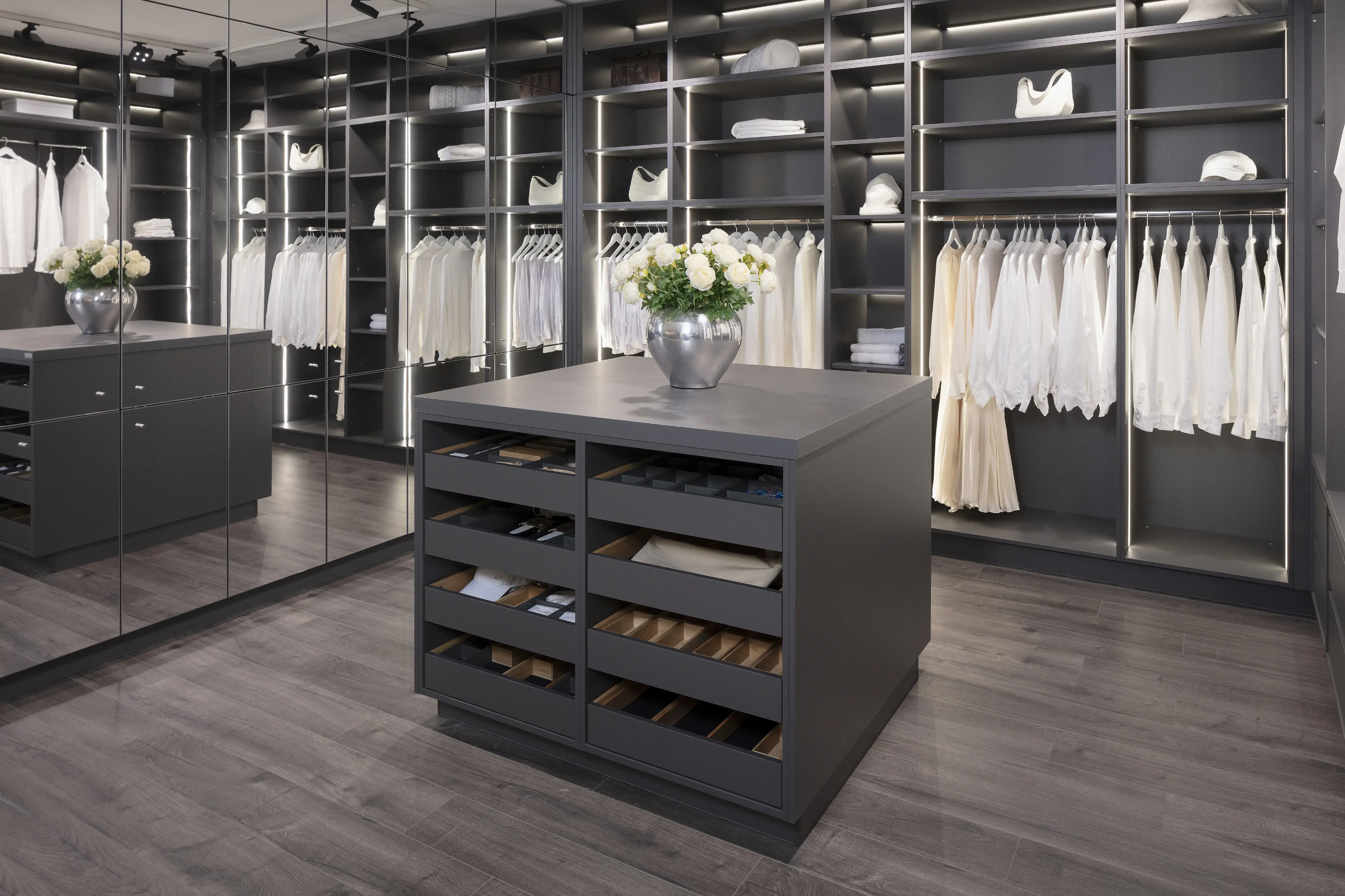 Ankleidezimmer von Ballerina mit Spiegelwand, Schrankwand mit Kleidung und einer Insel die Schubladen für Gürtel und Schuhe bietet. 