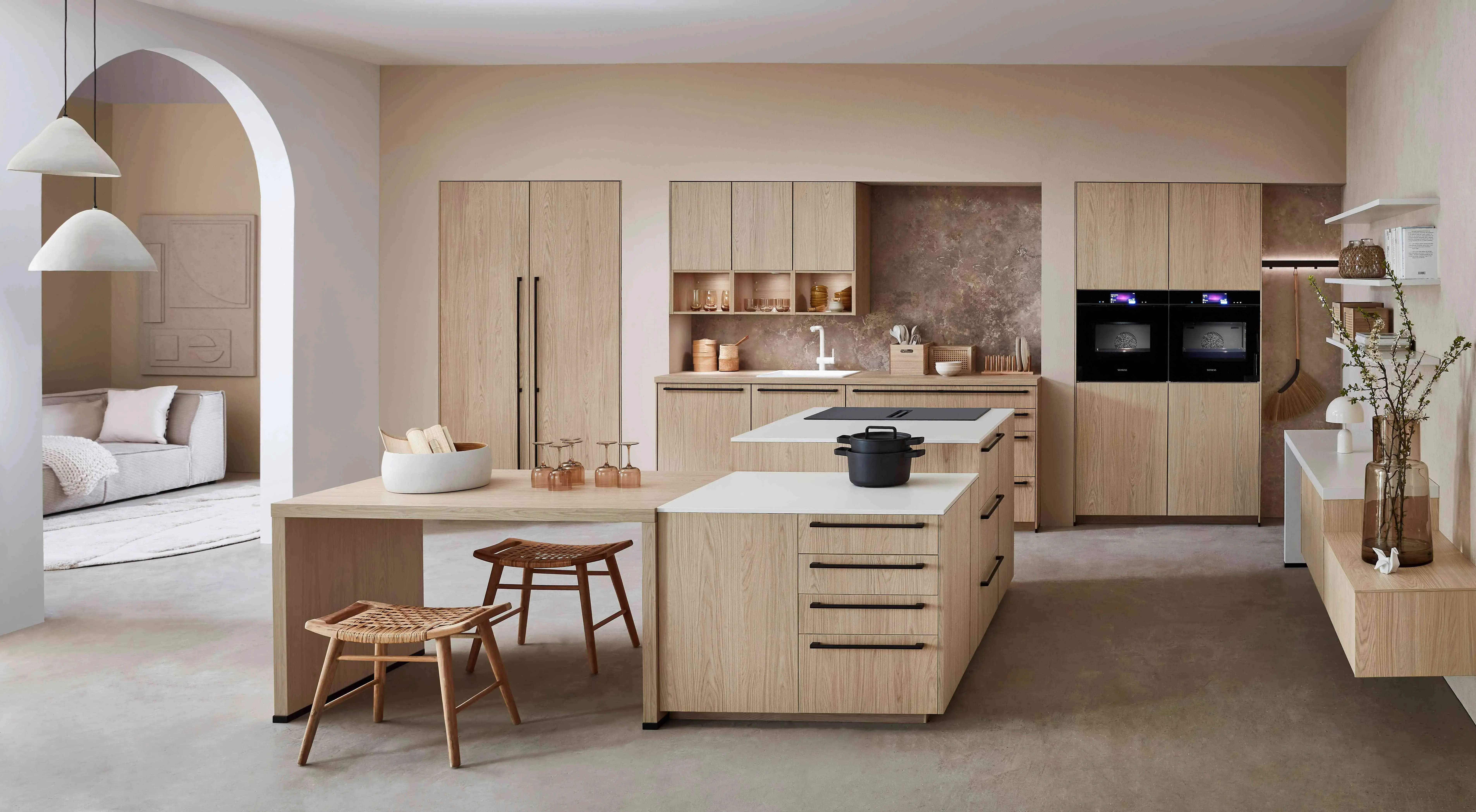 Hauptschuss, Mix aus Eichennachbildung und Premium Matt weiß, 2 Kücheninseln, eine mit Siemens Induktionskochfeld und Muldenlüfter, 2 Siemens Einbaubacköfen, Sitzbereich, 3 Oberschränke über der Spüle, Regalelement an der Wand, graue Nischenrückwand, Siemens Kühlschrank, Siemens Geschirrspüler